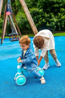 Older brother is helping his younger brother to ride a balance bike on a blue playground surface