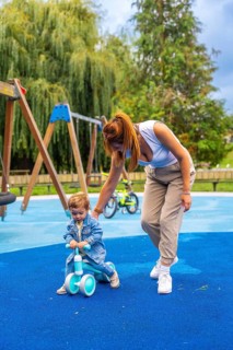 Young mother helping her toddler son learn to ride a balance bike on a playground's soft blue