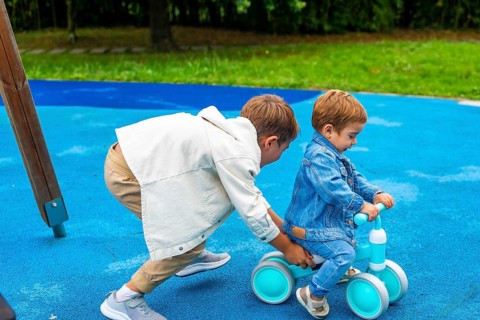 In a vibrant playground setting, an older brother supports his younger sibling as he learns to ride