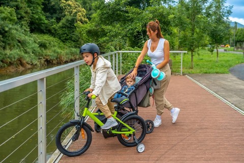 Young mother pushing a stroller with a baby inside while watching her other son riding a bicycle