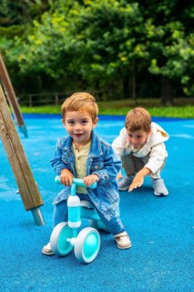 Happy toddler boy riding a balance bike on a playground with his older brother cheering him on,