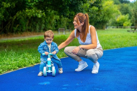 Young mother crouching next to her son helping him learn to ride a balance bike on a blue surface