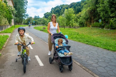 Mother pushing a stroller while walking alongside her son riding a bicycle in a vibrant park,