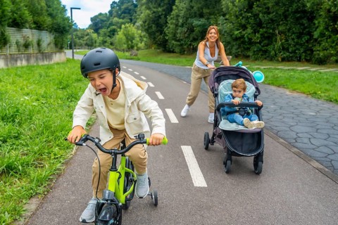Child wearing a helmet is riding a bicycle and shouting, while his mother is pushing a stroller