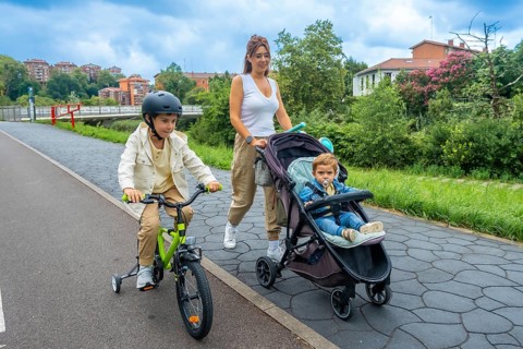 Mother enjoys a leisurely stroll in an urban park, pushing a stroller with her baby while her older