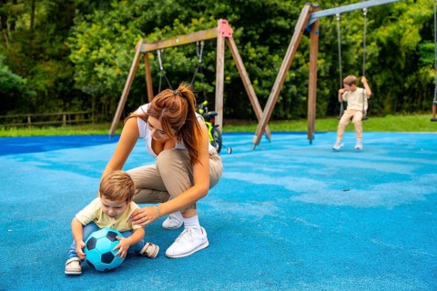 Young mother crouching next to her toddler son playing with a ball on a blue playground surface