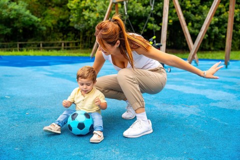 Young mother crouching beside her toddler son, guiding him as he plays with a soccer ball on a