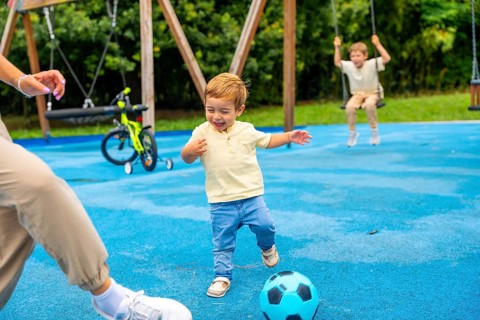 Happy toddler boy kicking soccer ball at playground with family, enjoying a playful moment on a