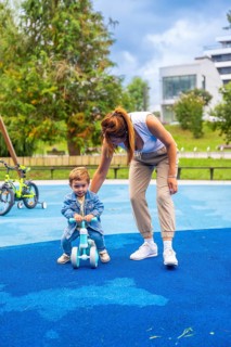 Young mother guiding her son as he learns to ride a balance bike on a playground featuring a soft