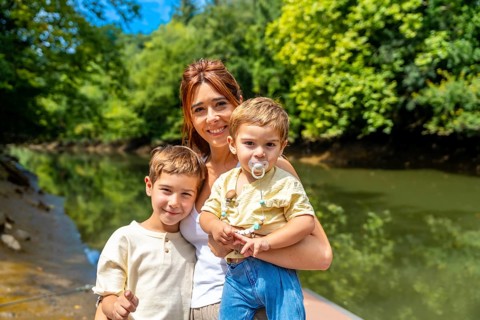 Happy mother holding her baby and posing with her older son by the river in a sunny summer day,