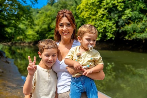 Caucasian mother joyfully holding her two sons near a river on a sunny summer day, with one son