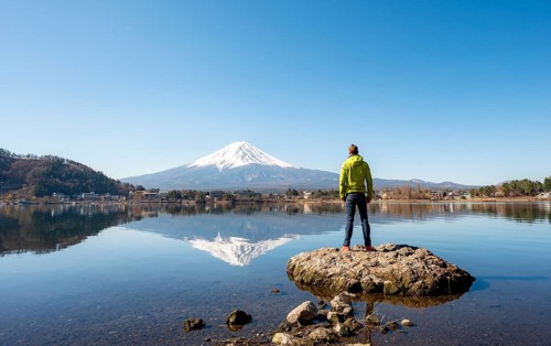 Young man standing on a rock in water, volcano Mt. Fuji is reflected in Lake Kawaguchi, Yamanashi