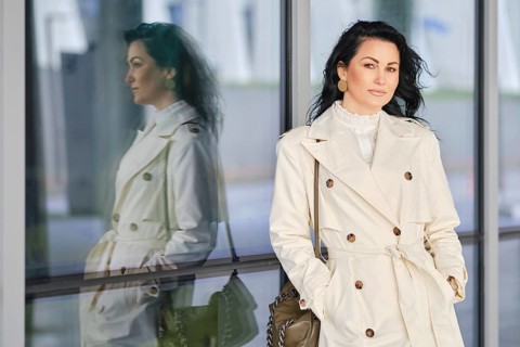 A woman dressed in a chic trench coat stands confidently beside a reflective glass wall. Her long
