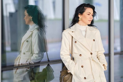 A woman dressed in a fashionable trench coat stands outside, her reflection visible in the glass