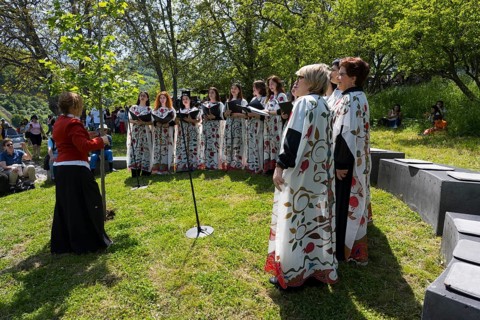 Women's choir in white colorful robes singing outdoors, surrounded by trees and audience, choir at