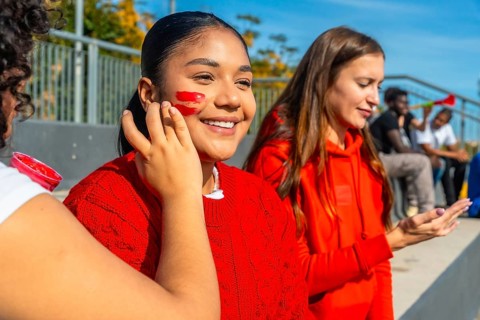 Young happy spectators sitting in bleachers having face paint applied before a sports event,