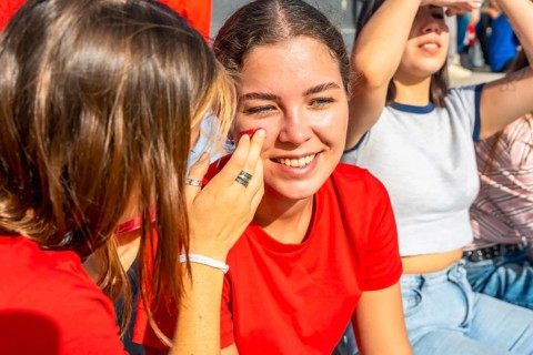 Young women at a sunny stadium, one friend painting red face paint on anothers cheek as they smile