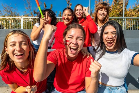 Diverse group of young women cheering loudly in a stadium, faces painted and fists raised,