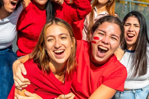 Group of excited young women wearing red shirts and face paint, screaming and cheering while