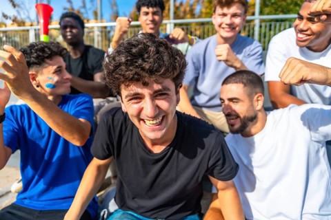 Young diverse male friends cheering in a sunlit stadium, celebrating their team with smiles,