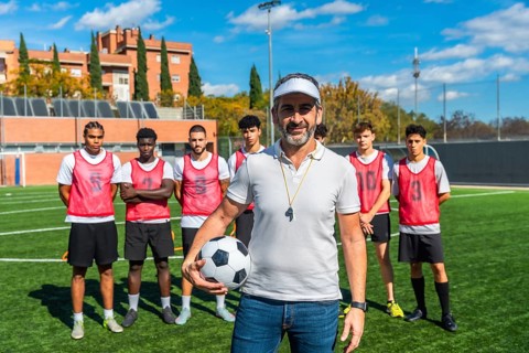 Male coach with a confident smile standing on a football field, holding a soccer ball, with his