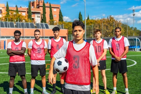 Diverse group of male soccer players in red bibs standing on a training pitch, holding a ball and