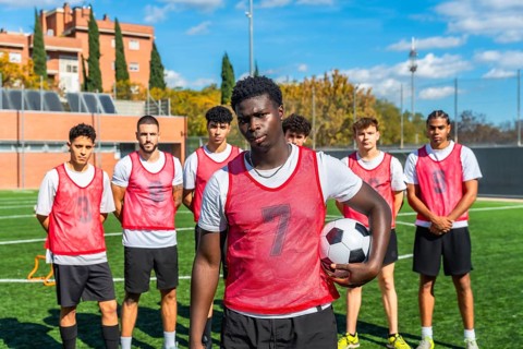 Young multi ethnic men's soccer team in red bibs and white shirts standing on a sunny green pitch,