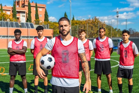Soccer captain stands with ball on green pitch, leading a diverse, determined team of young male