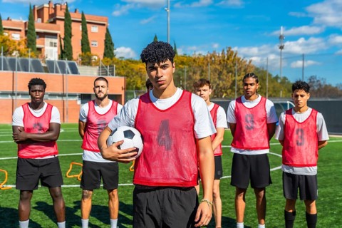 Multi ethnic group of young men in red bibs standing on a green football pitch, preparing for
