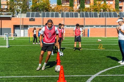 Young men in red vests are performing agility drills with cones and soccer balls on a green
