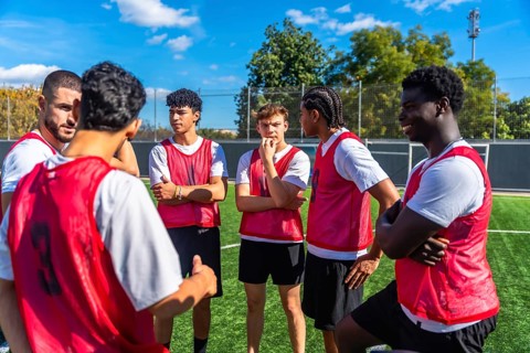 Young male soccer players wearing red bibs huddling on a green artificial turf field, listening