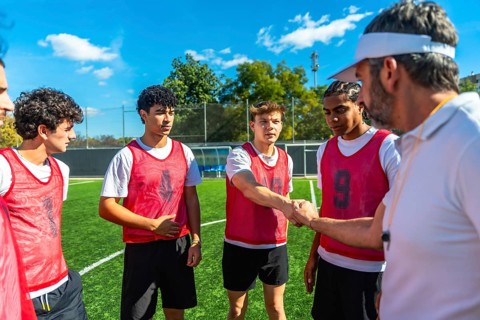 Coach wearing a visor and whistle shaking hands with a young male soccer player on a green football
