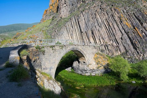 An old stone bridge stands in front of dramatic rock walls surrounded by green vegetation and a
