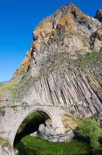 A high stone bridge stretches in front of an impressive rock formation under a bright blue sky,