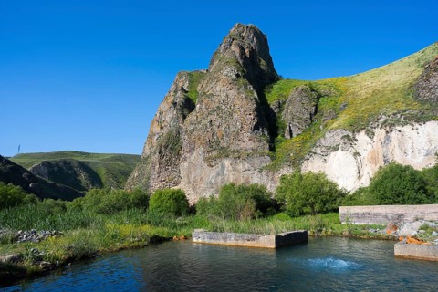 Clear water with spring flows through a lush landscape dominated by huge, rocky mountains, hot