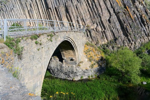 View of a stone bridge across a clear river with lush greenery and dramatic rock formations in the