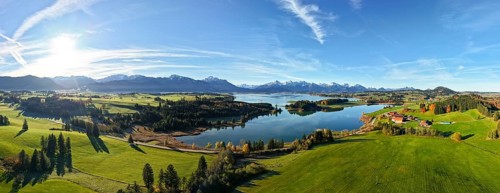 Wide panoramic view of a green landscape with lake and mountain backdrop under clear sky,