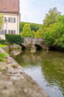 An old stone bridge over a quiet river flanked by buildings and trees, ZFP Zwiefalten, Reutlingen