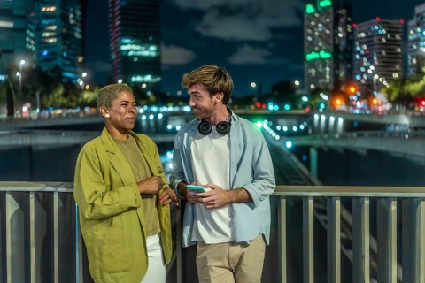 Diverse young couple chatting and smiling on an urban bridge at night, city skyline and glowing