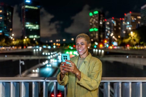 Woman standing on a city bridge at night, smiling while using a smartphone with blurred traffic and