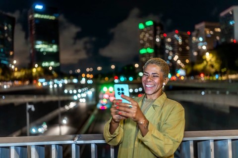 Happy woman smiling, holding a smartphone and enjoying a vibrant urban night view from a bridge,