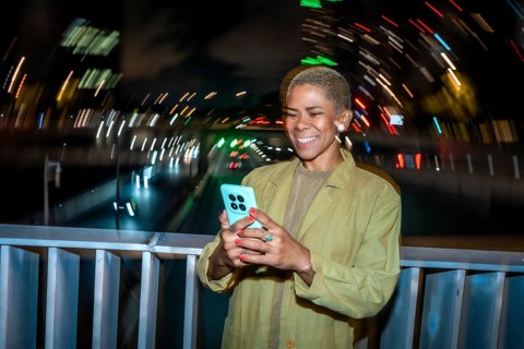 Smiling young adult woman using a mobile phone at night on a city bridge, enjoying digital
