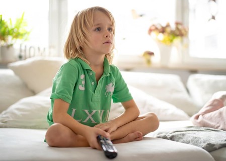 Child, boy, 7 years old, blonde, sitting on sofa, with remote control in hand, watching TV,