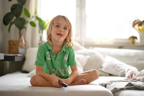 Child, boy, 7 years old, blonde, sitting on sofa, with remote control in hand, watching TV,