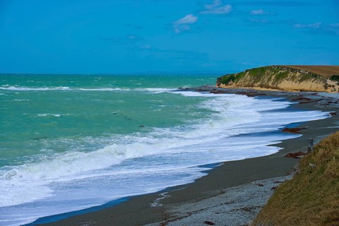 Waves crash against a rocky coast with blue sky, Tuhawaiki Point Lighthouse or Jack's Point