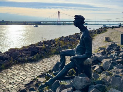 Photo with melancholy mood of bronze sculpture Poortekerl looking at Rhine waiting for ship to