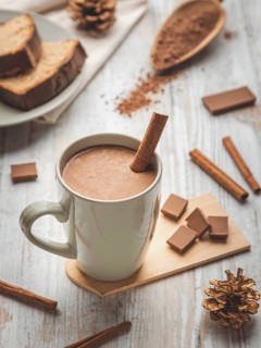 A cup of hot chocolate with cinnamon stick surrounded by cocoa powder and cake