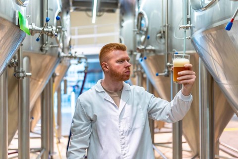 Brewmaster in lab coat inspects a glass of golden craft beer in a modern brewery, assessing color,