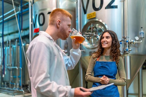 Brewery team performs quality control tasting and discussion beside stainless steel fermenters,