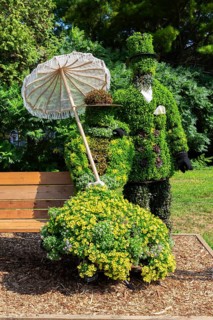 Live plant sculpture of Victorian man standing and woman with sun shade umbrella sitting on wooden
